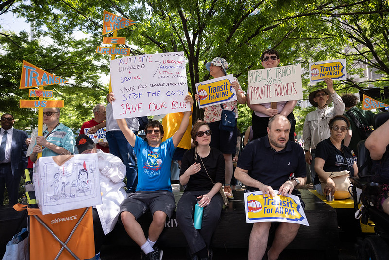 A crowd of people at a rally holding signs that say "transit for all PA!" and other slogans about saving bus service.