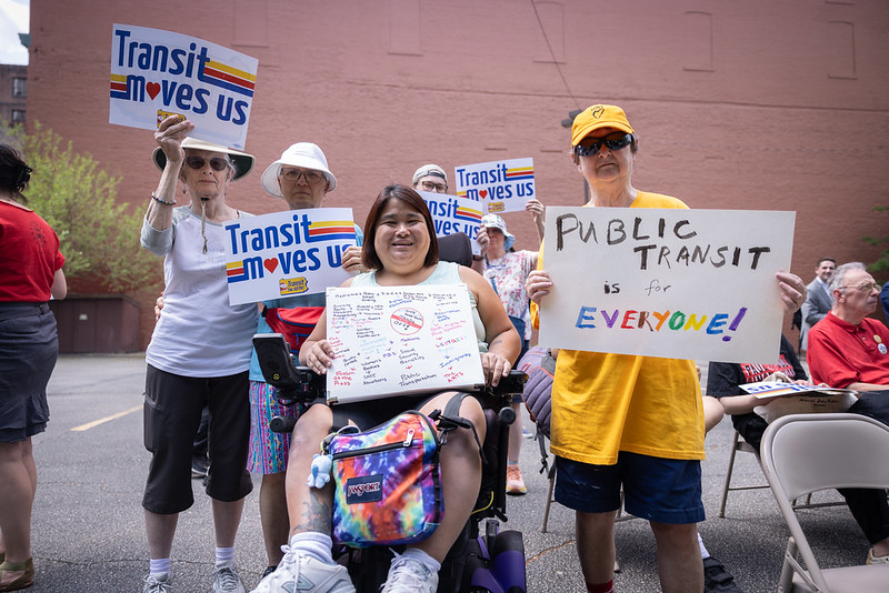 A group of people hold signs that say "transit moves us", "public transit for everyone", and other slogans.