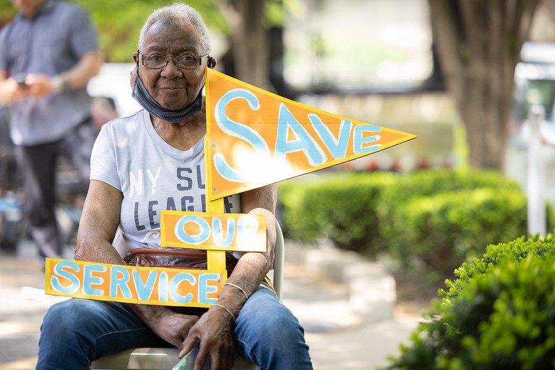 A Black woman holds a colorful orange and blue handmade sign reading "save our service"
