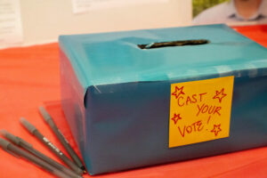 A blue ballot box for our 2026 Board of Directors, decorated with red and yellow "cast your vote!" and stars