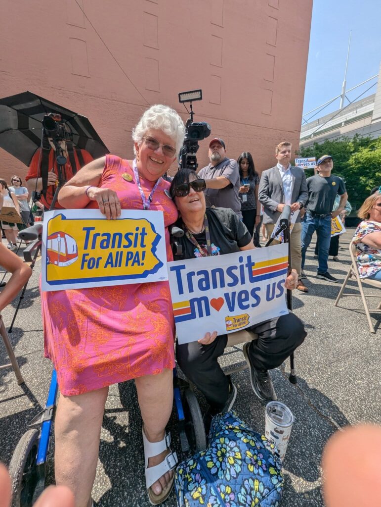Two transit advocates sit at a rally on chairs and rollators, holding red, yellow, and blue "Transit for All PA!" and "Transit moves us" signs.