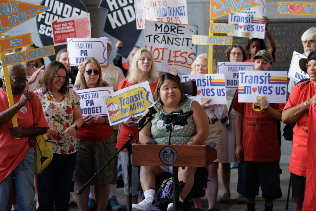 A crowd of transit advocates holds colorful signs at a rally behind a woman in a wheelchair, speaking into a mic at a wooden podium.