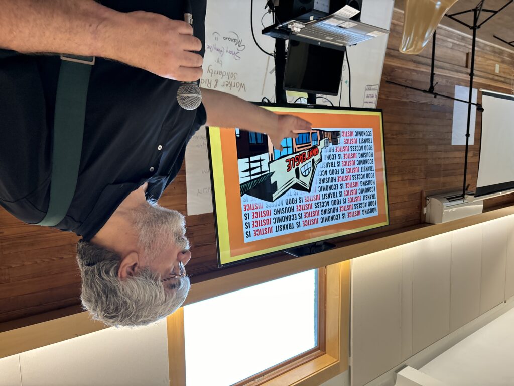 A light-skinned, gray-haired man speaks into a mic, gesturing at an illustration of a grocery store reading "housing justice is economic justice is transit justice is..."