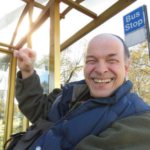 Bill McDowell, a white man raising his fist and smiling at a sunny PRT bus stop