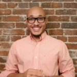 Kristopher Chandler, a Black man smiling in a pink shirt against a brick wall